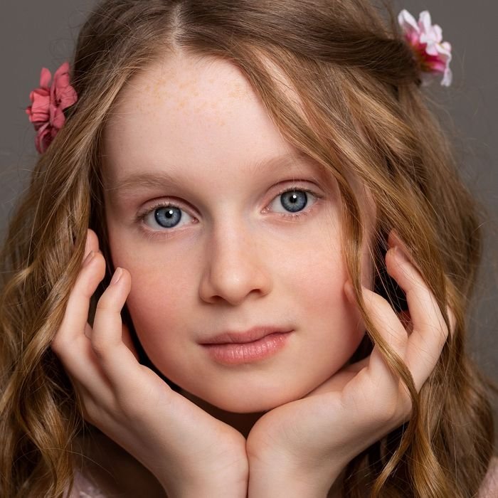 Charming close-up portrait of a young girl with flowers in her hair, hands framing her face.